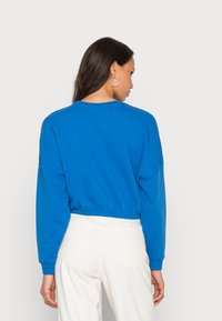 Sudadera recortada de color azul royal con mangas largas y cuello redondo. Tejido texturizado, dobladillo elástico y mangas con puños. Combinada con pantalones de color claro.