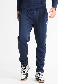 Marineblå træningsbukser med elastisk talje, glat tekstur og sidelommer; parret med sorte og hvide sneakers.