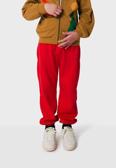 Rote Trainingshose, braune Zip-Jacke mit bunten abstrakten Mustern und weiße Sneakers. Die Jacke hat ein zweifarbiges Design.