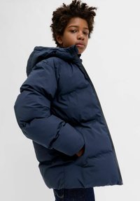 Tumši zils pūkains jaka ar kapuci, aprīkota ar rāvējslēdzēju un sānu kabatām. Izgatavota no gluda, izolējoša materiāla ar pufainu tekstūru.