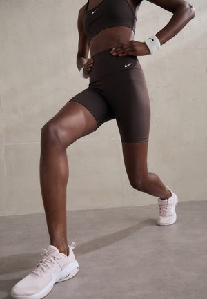 Athletische Person trägt schwarzen Nike Sport-BH und Shorts, weiße Sneakers und Handgelenksbänder, in einer Ausfallschritt-Übungs-Pose vor neutralem Hintergrund.