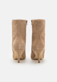 Ett par beige mockaläder ankelskottar med smala högklackar och bakdragkedjor, visade bakifrån på en vit bakgrund.