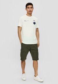 Biała bawełniana koszulka z granatowymi grafikami, w połączeniu z oliwkowymi szortami cargo z wieloma kieszeniami, oraz białymi sneakersami.