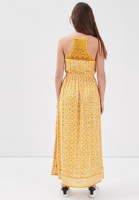 Robe maxi avec un motif floral jaune et blanc. Elle possède un dos en crochet texturé et une taille élastique. Portée avec des baskets blanches et noires.