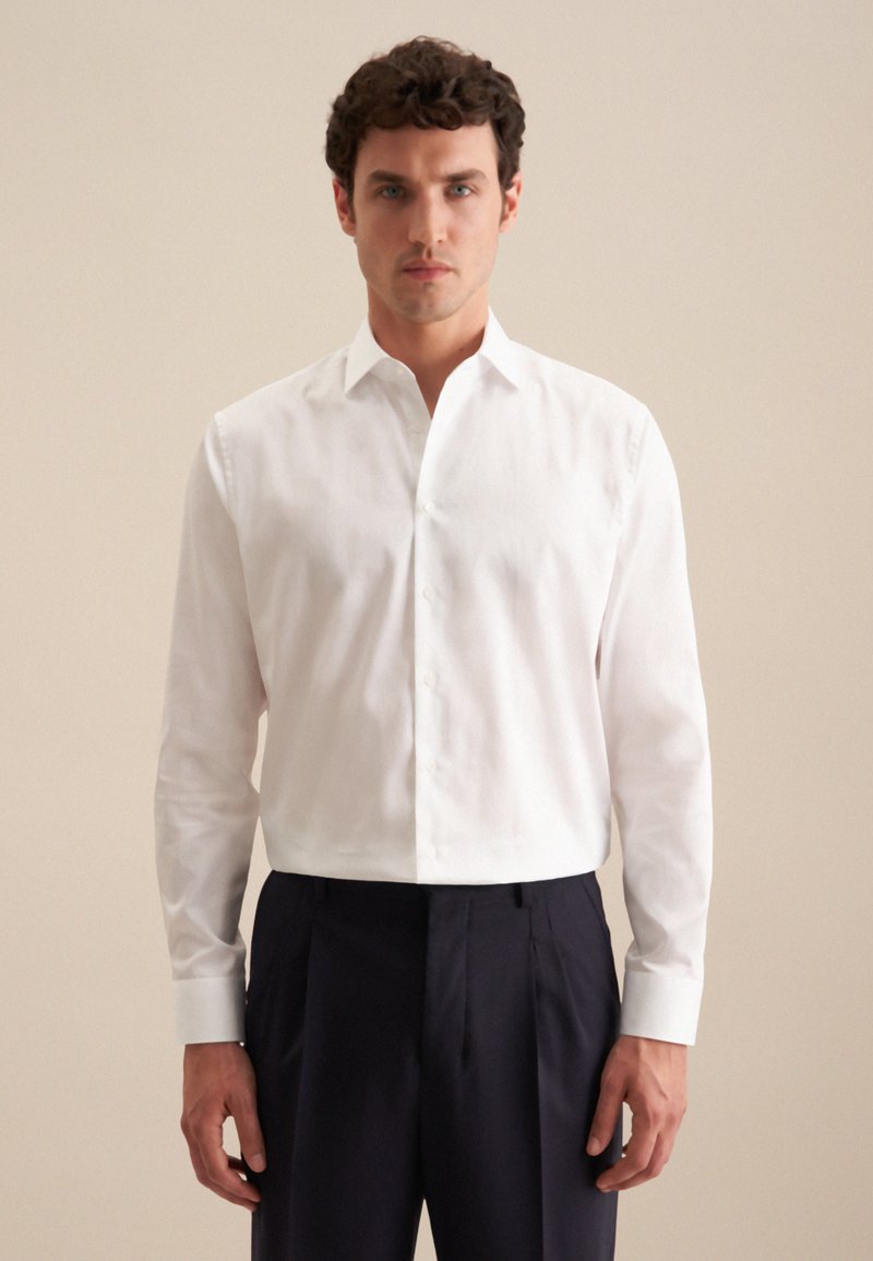 Witte button-up shirt van gladde katoen; heeft lange mouwen en een klassieke kraag. Gepaarde met donkere geplooide broek.