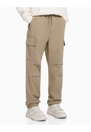 Person, der beigen Cargo-Hosen mit mehreren Taschen und weißen Sneakers trägt, mit Händen in den Taschen, im Stehen vor einem weißen Hintergrund.