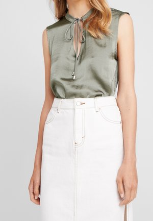 Mouwloze olijfgroene satijnen blouse met striksluiting bij de hals, gecombineerd met een witte high-waisted denim rok met een voorste split.