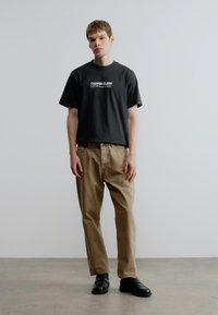 Melna kokvilnas T-krekls ar "CALVIN KLEIN NEW YORK" grafiku, kopā ar gaiši bēšām biksēm, valkāts ar melnām kurpēm uz gludas virsmas.