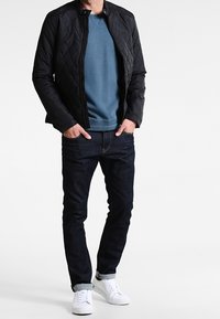 Sort quiltet jakke over en blå crewneck sweatshirt, parret med mørke jeans og hvide sneakers. Outfitet har en afslappet stil.