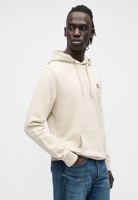 Felpa con cappuccio beige con tasca frontale, coulisse regolabili e un piccolo logo giallo. Modello abbinato a jeans blu. Tessuto liscio.