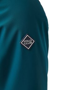 Teal atletisk t-shirt gjord av slät tyg, med en diamantformad "POLO CLUB" logopatch på vänster axel.