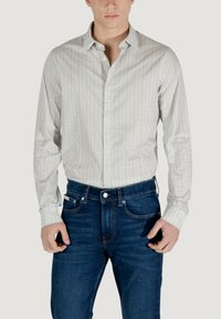 Camicia bianca a maniche lunghe con un sottile motivo geometrico, colletto button-down, abbinata a jeans di denim blu scuro. Texture liscia, design aderente.
