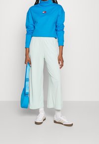 Blaues Sweatshirt mit hohem Kragen und Logodetail, kombiniert mit hellgrünen weit geschnittenen Hosen. Weiße Sneaker mit beigen Akzenten und einer blauen Tasche.