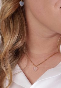 Gouden ketting met een doorschijnende harthanger, vergezeld van hartvormige oorbellen. De achtergrond toont een deel van een wit kledingstuk.