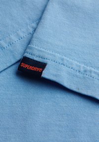 Lichtblauw T-shirt met een gladde textuur en korte mouwen, met een zwart label waarop "SUPERDRY" in het rood staat.