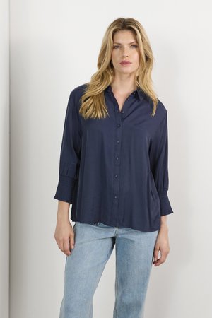 Chemise bleu marine à col avec des boutons, à manches longues et smockées, avec une coupe fluide. Tissu lisse avec un léger éclat, associé à un jean bleu clair.