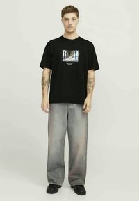 Melnas kokvilnas T-krekls ar palmu grafiku, apvienots ar izbalējušām pelēkām platām biksēm un melnām kurpēm. Modelis atrodas neitrālā stāvoklī.