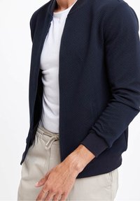 Granatowa bomberka z teksturowanym wzorem, zamkiem błyskawicznym i ściągaczami przy mankietach, noszona na białej koszulce i beżowych spodniach ze sznurkiem w pasie.