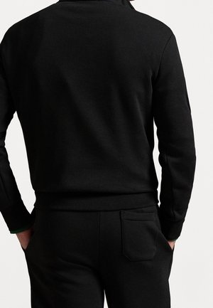 Mann trägt ein eng anliegendes schwarzes Langarmshirt und schwarze Hose mit Gesäßtasche, Hände in den Taschen, von hinten vor weißem Hintergrund gezeigt.