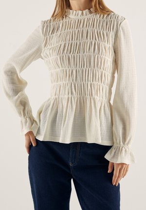 Femme portant un chemisier texturé crème à manches longues avec des détails élastiques froncés et des poignets à volants, associé à un jean bleu foncé.