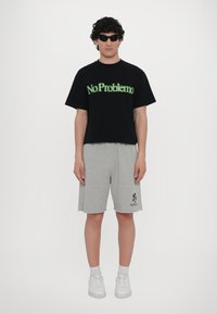 Tricou din bumbac negru cu grafică verde "No Problemo", asortat cu pantaloni scurți din bumbac gri cu un logo și adidași albi. Păr creț și ochelari de soare.