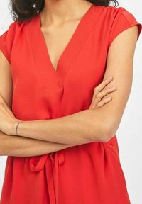 Blouse rouge à manches courtes avec un décolleté en V, détail à nouer à la taille et texture lisse. Design simple sans motifs ni embellissements.