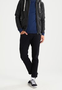 Grauer Reißverschluss-Hoodie mit weißen Kordelzügen, blauer Pullover, schwarze Hose und schwarze Sneaker mit weißen Akzenten sowie Socken. Einfaches, lässiges Design.