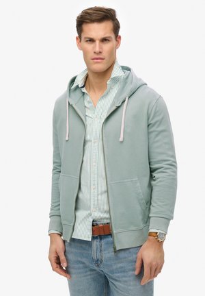 Sudadera verde claro con cremallera y capucha ajustable, bolsillos frontales y puños acanalados, superpuesta sobre una camisa de rayas azul clara con botones.