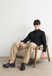 Zwarte overhemd met knopen en wit logo, beige broek en zwarte instappers. Model draagt een donkere zonnebril en zit op een lichtgekleurde fauteuil.