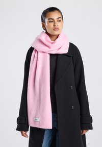 Sciarpa di lana rosa appesa attorno al collo, caratterizzata da una texture morbida. Indossata con un cappotto doppiopetto nero, con un'etichetta sul bordo della sciarpa.