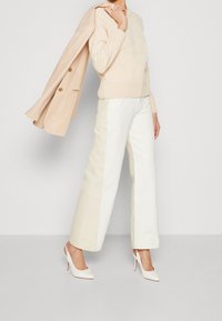 Kvinna klädd i en krämfärgad kabelstickad tröja och tvåtoniga vida byxor, som håller en beige kavaj över axeln, med vita högklackade skor.