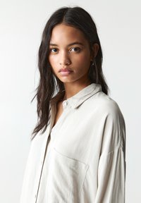 Chemise boutonnée beige clair avec un col et une poche sur la poitrine. Coupe ample, manches longues, texture lisse, adaptée pour une tenue décontractée ou semi-formelle.