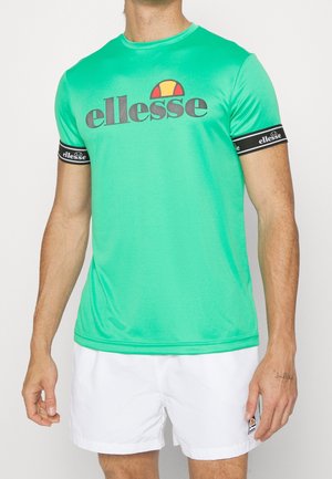 Mand iført en grøn ellesse t-shirt med sorte logobånd på ærmerne og hvide ellesse shorts.