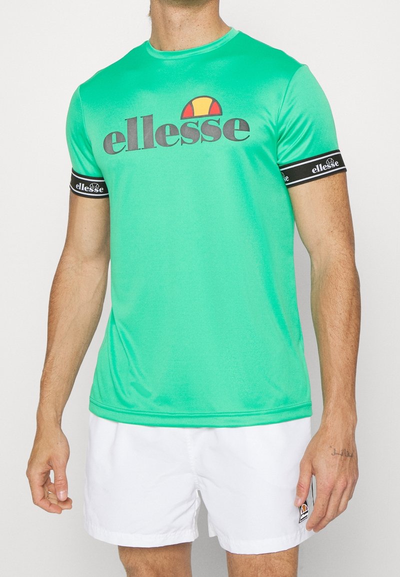 Homme portant un t-shirt ellesse vert avec des bandes noires avec le logo sur les manches et un short ellesse blanc.