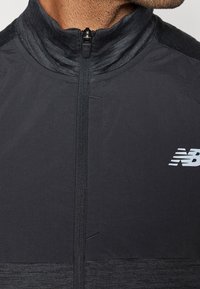 Close-up of a partially zipped black New Balance jacket with a high collar worn by a person.