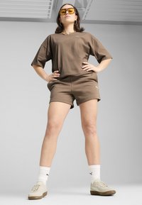 Ensemble en coton marron, composé d'un t-shirt et d'un short, offrant une coupe décontractée. Les accessoires incluent des lunettes de soleil jaunes et des chaussettes blanches avec un logo noir, le tout associé à des baskets beiges.