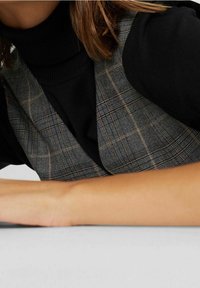 Zwarte coltrui onder een grijs geruit vesttopje met dunne bandjes, voorzien van lichtbruine accenten. Glad oppervlak, close-up van stofdetails.