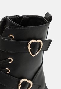 Bottine noire en cuir synthétique avec un bout rond, accents en métal doré en forme de cœur et détails en dentelle. Présente une texture lisse et une conception robuste.
