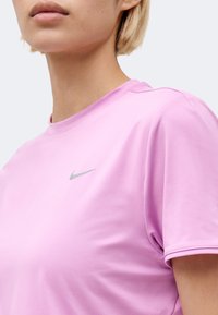 Κοντινό πλάνο ενός ατόμου που φοράει ένα ανοιχτό μοβ μπλουζάκι Nike με μικρό ασημένιο λογότυπο Nike στο στήθος, ορατό από τους ώμους μέχρι το πηγούνι.