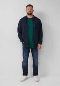 Marine blauwe zip-up hoodie over een groene t-shirt, gecombineerd met donkere jeans en witte sneakers. Soepel materiaal, casual pasvorm, eenvoudige stijl.