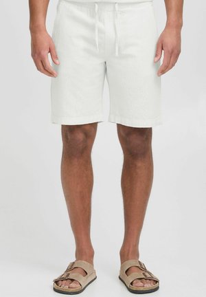 Lysfarvede linnedshorts med elastisk taljebånd og snøre. Knælange med kantede manchetter; båret med beige sandaler.