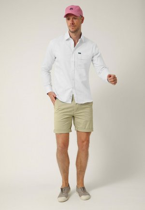 Hombre con una camisa blanca abotonada, pantalones cortos beige remangados, zapatos grises sin cordones y una gorra de béisbol rosa, de pie frente a un fondo liso.