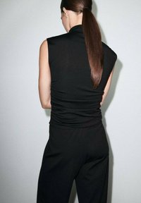 Donna con una lunga coda di cavallo scura, vestita con un top senza maniche nero e pantaloni neri, rivolta verso un muro chiaro e semplice.