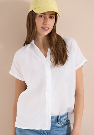 Ung kvinde med langt brunt hår iført en gul kasket, hvid kortærmet skjorte med knapper og lyseblå jeans mod en beige baggrund.