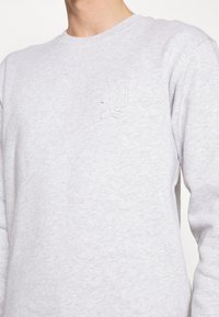 Sweat-shirt gris clair avec une encolure ronde et un discret logo brodé "D" sur le côté gauche de la poitrine. Le tissu semble doux et texturé.