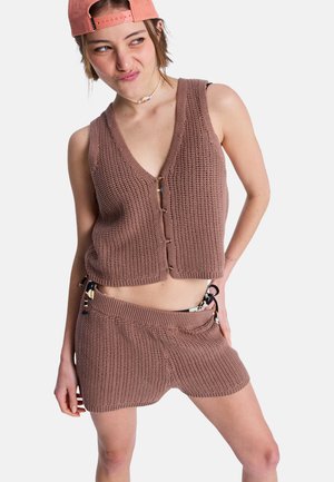 Młoda osoba ubrana w brązową, dzianą kamizelkę bez rękawów i pasujące szorty, z różową czapką założoną do tyłu i naszyjnikiem z koralików.