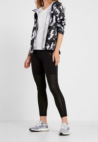 Chaqueta de patrón en blanco y negro, camiseta blanca, leggins negros con paneles translúcidos y zapatillas con patrón con acentos grises y azules.