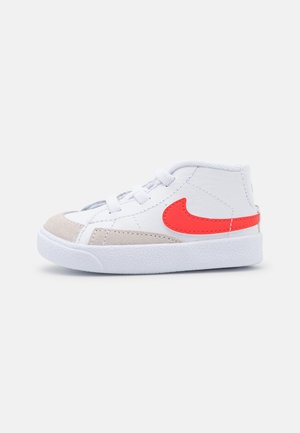 Basket montante blanche avec bout en daim beige et logo Nike Swoosh rouge vif sur le côté, vue de l'extérieur.