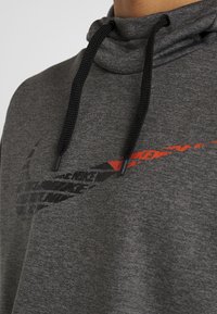 Haut à capuche gris avec des cordons noirs, présentant un tissu texturé et un logo Nike noir et rouge bien visible imprimé sur le devant.
