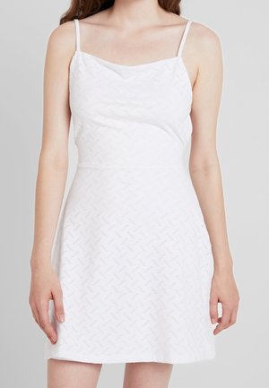 Femme portant une mini-robe blanche sans manches avec de fines bretelles et un subtil motif en zigzag, debout les bras détendus le long du corps.
